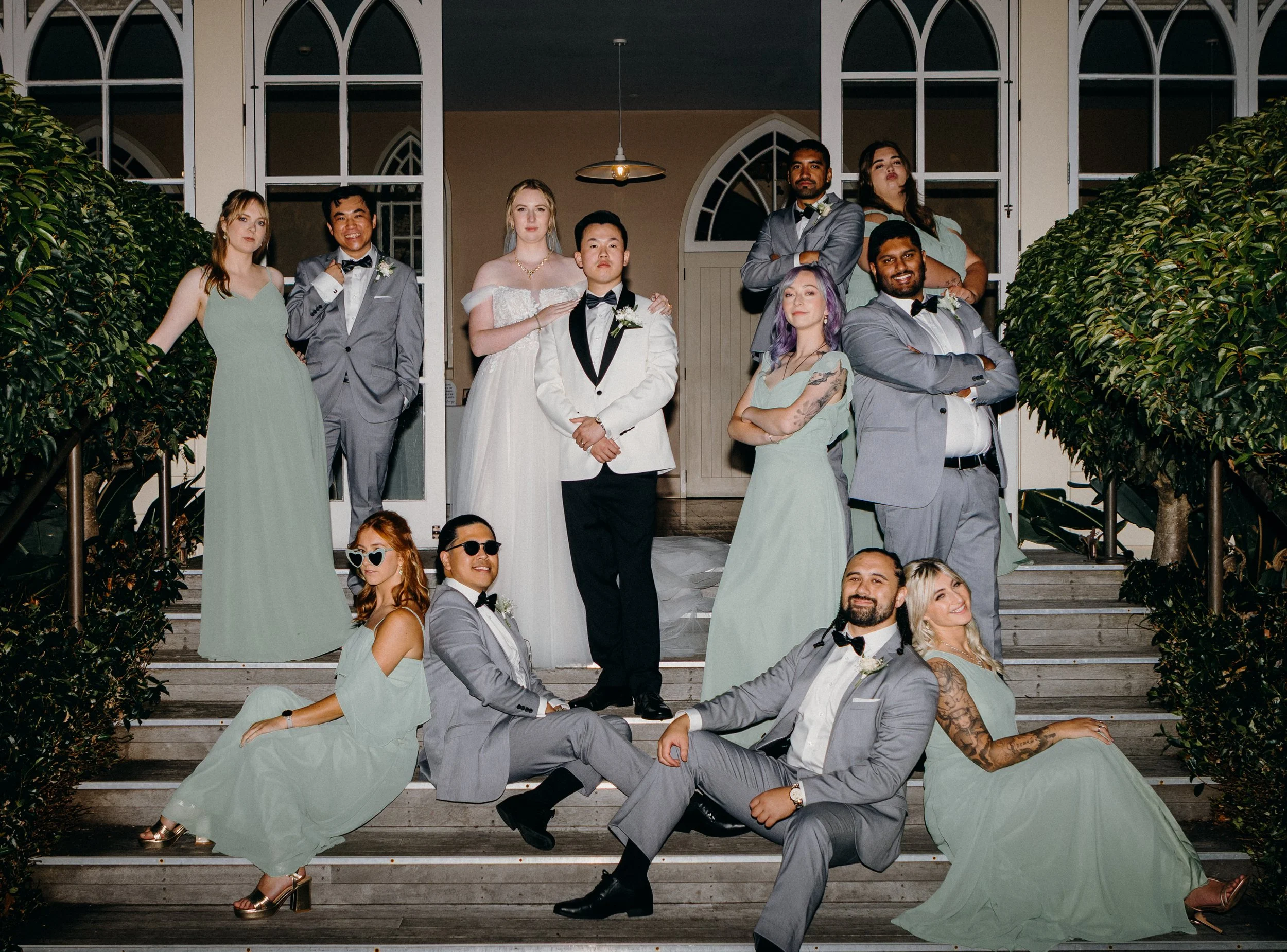 A group of people at a wedding, posing on a staircase in front of a building with glass doors and arched windows, dressed in formal and semi-formal attire.
