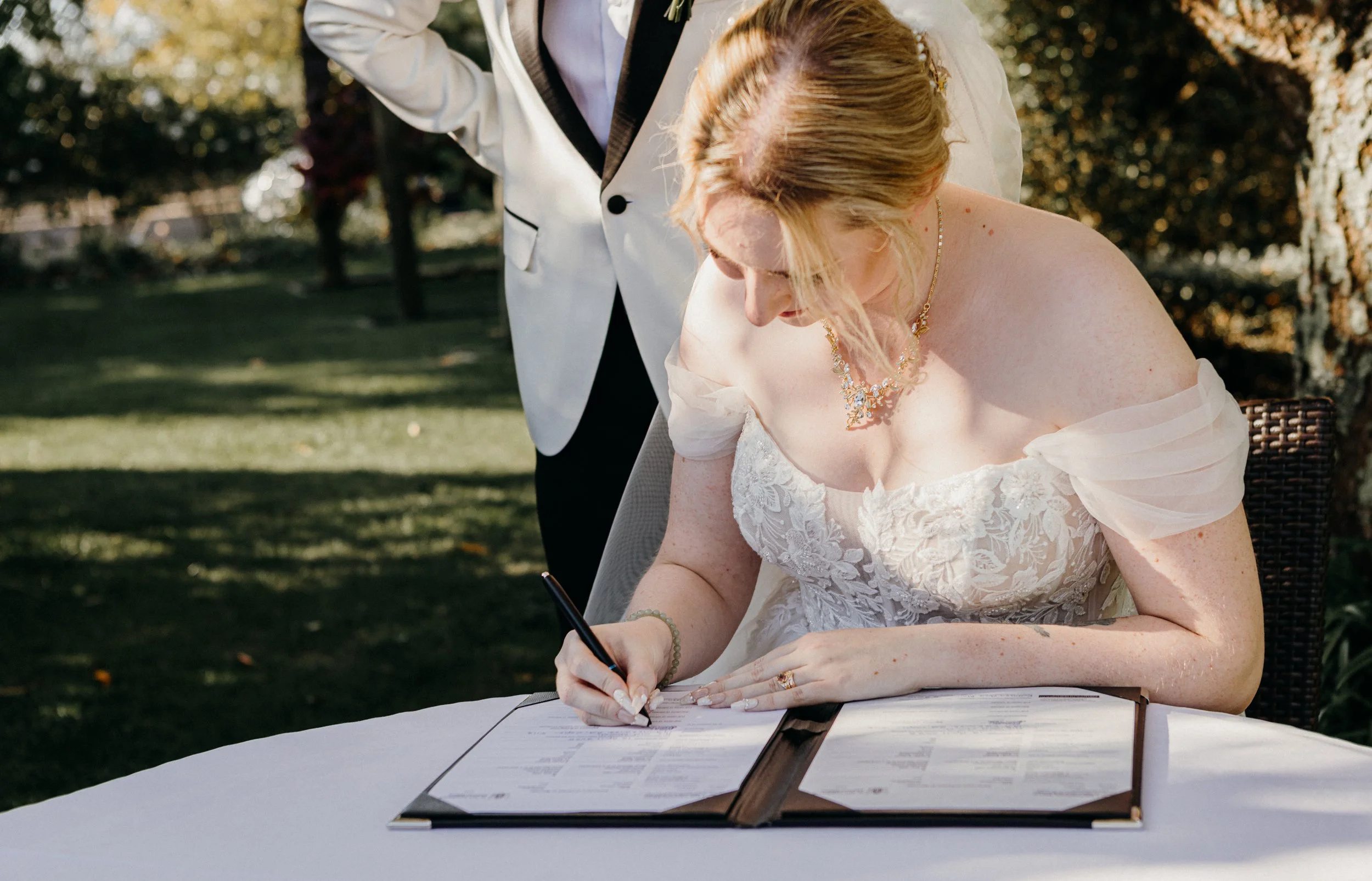 A bride signing a marriage certificate outdoor during daytime, wearing a white off-shoulder wedding gown with lace details, gold jewelry, and her hair styled in an updo, while a person in a white blazer stands beside her.