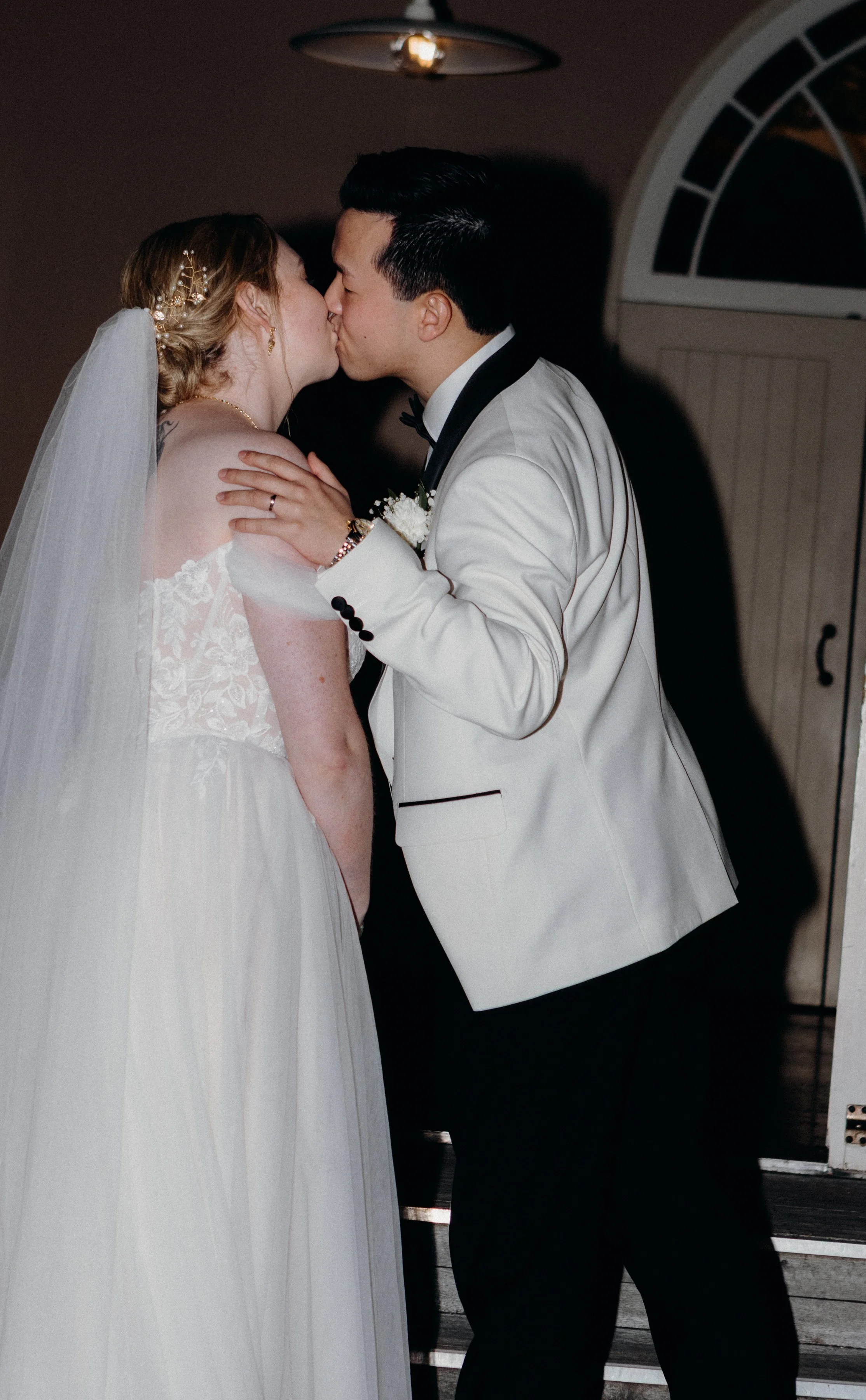 A bride and groom sharing a kiss at their wedding, with the groom in a tuxedo and the bride in a wedding dress and veil, inside a warmly lit room.