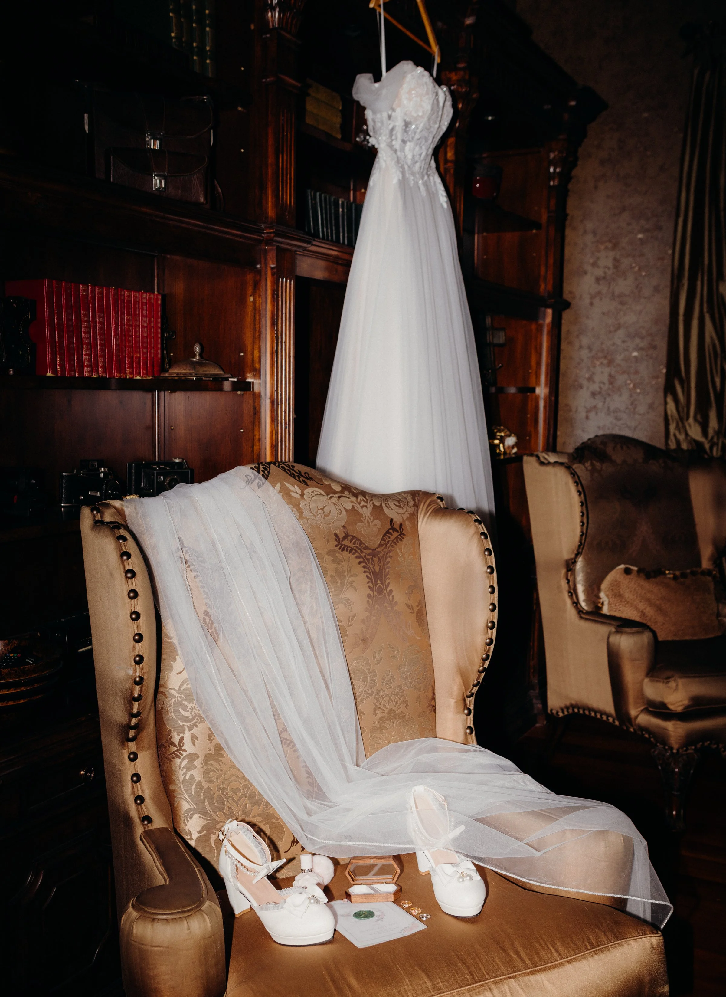 A wedding dress hangs from a yellow hook, with a pair of white bridal shoes with bows and a veil on a vintage upholstered armchair in a dimly lit room.