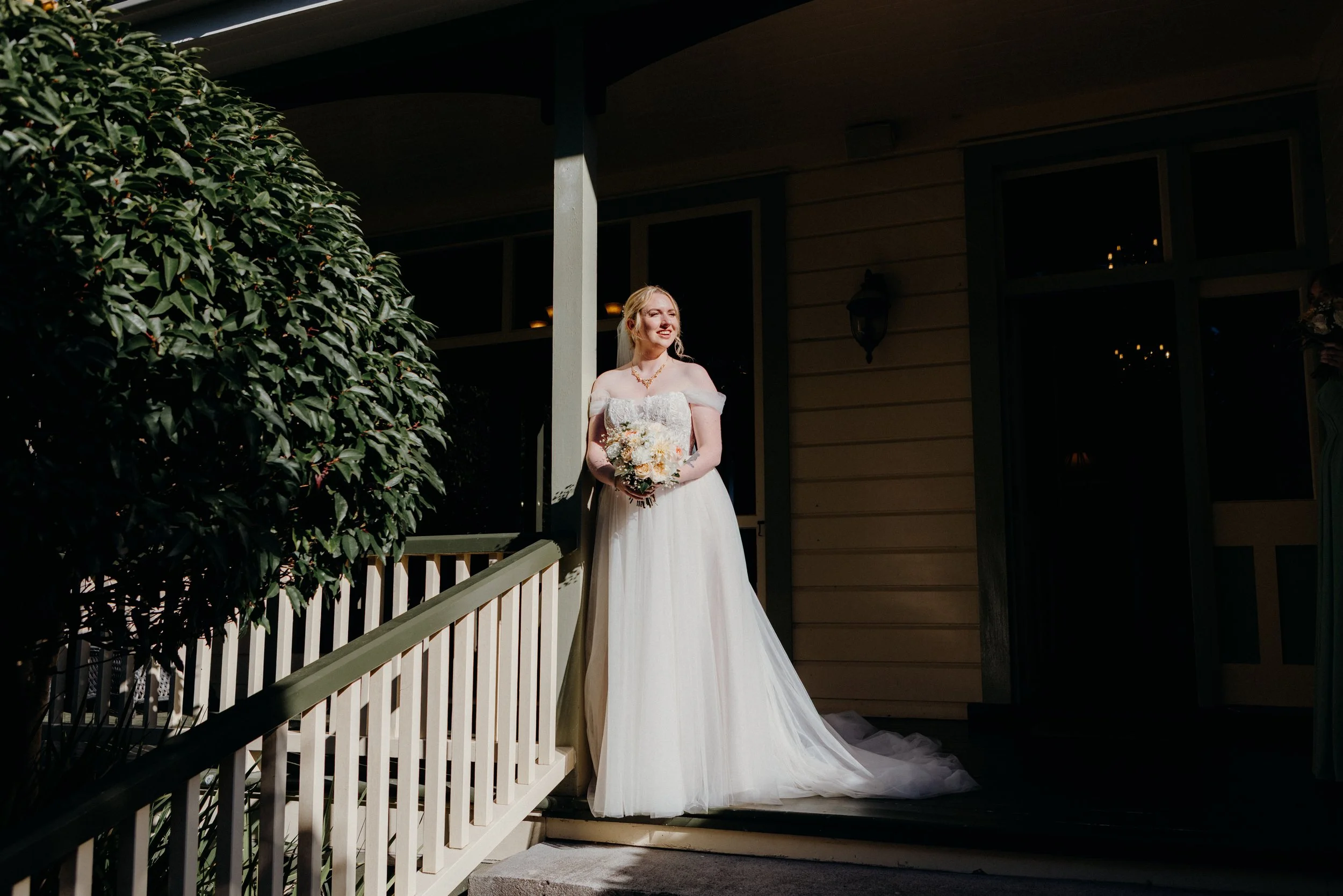 A bride in a white wedding dress holding a bouquet standing on a porch, illuminated by sunlight, with greenery on the left and a house in the background.