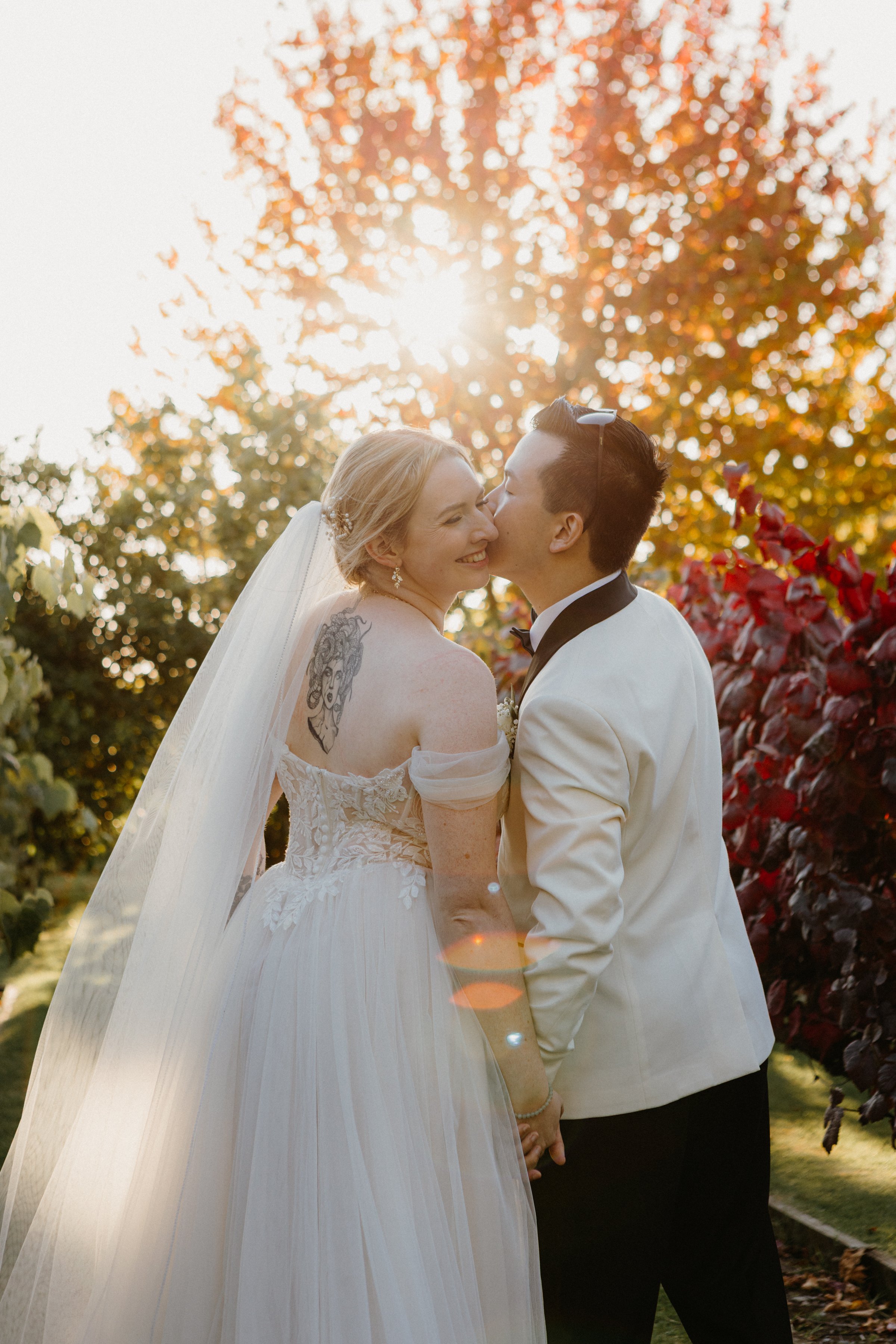 A newlywed couple, the bride with blonde hair in an off-shoulder wedding dress showing a tattoo on her shoulder, and the groom in a white tuxedo, sharing a kiss outdoors during sunset amidst autumn trees.