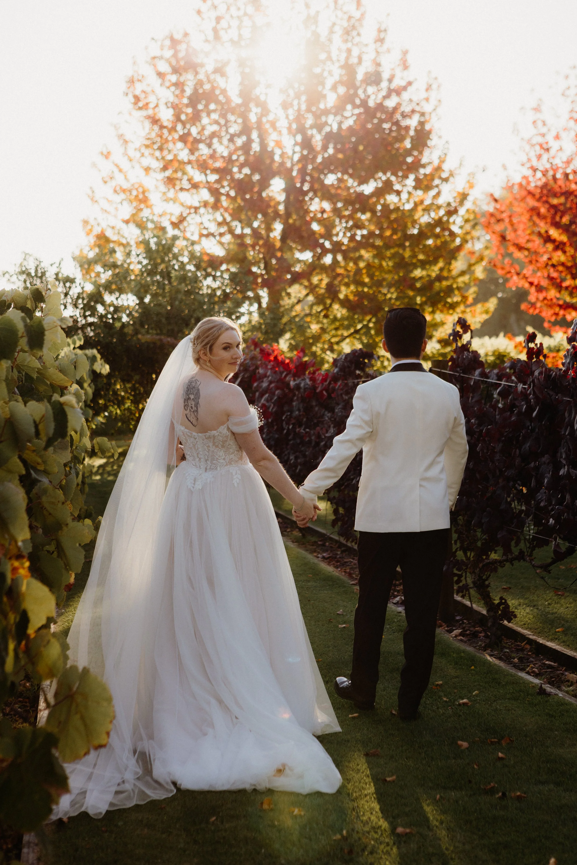 A bride and groom holding hands walking through a vineyard on a sunny autumn day, with the bride wearing a white lace wedding gown and veil, and the groom in a white tuxedo jacket and black pants.