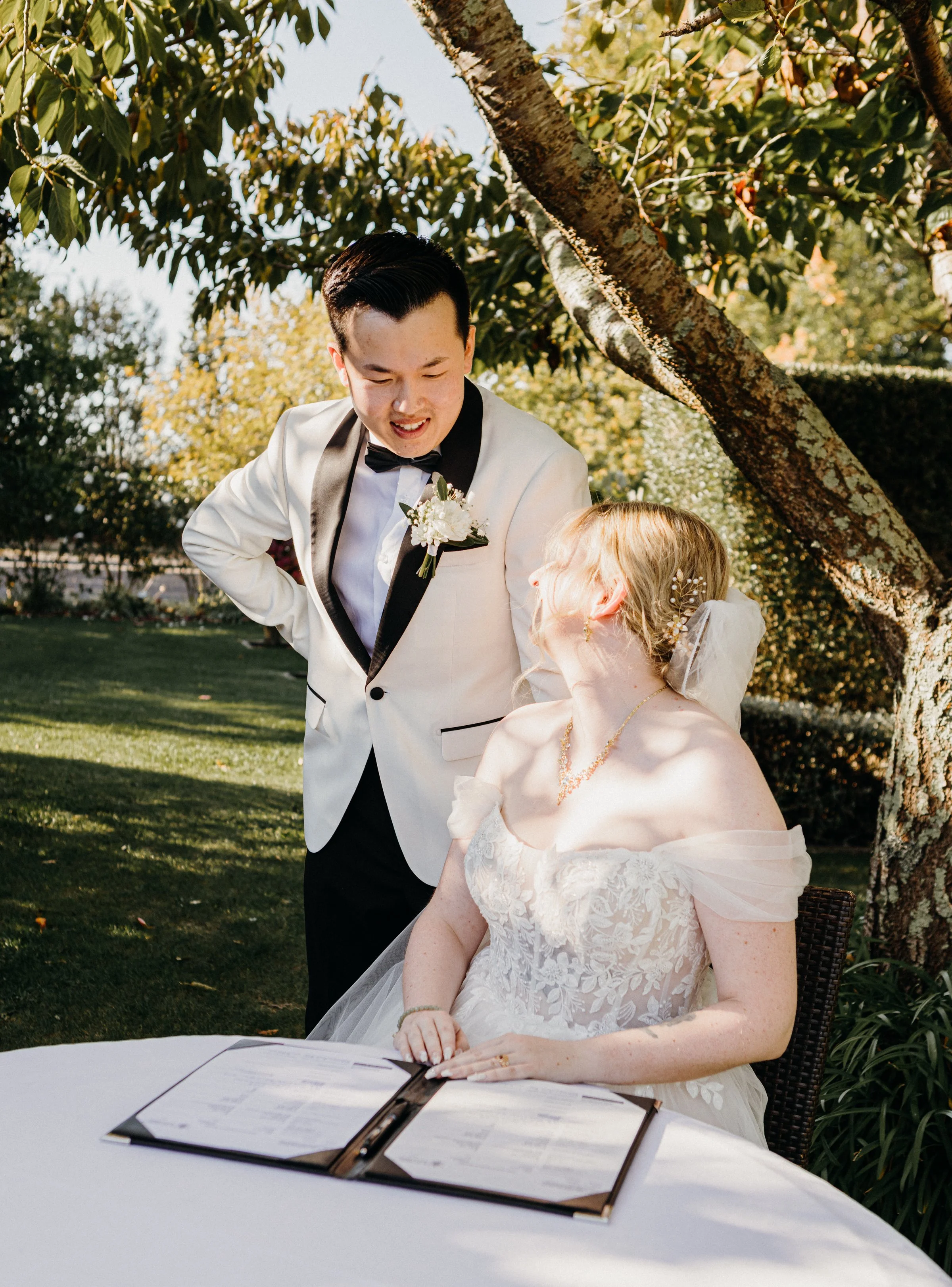 A newlywed couple signing their marriage license outdoors, the groom leaning over to talk to the bride, under a tree with sunlight filtering through the leaves.