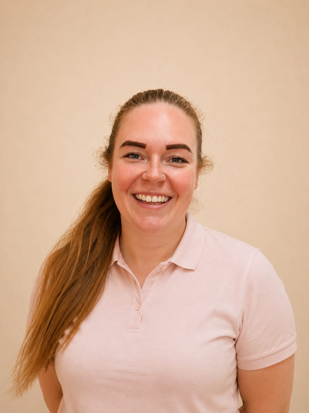 Lachende vrouw met lang rood haar, donkerbruine wenkbrauwen, in een lichtroze polo, staand voor een beige muur.