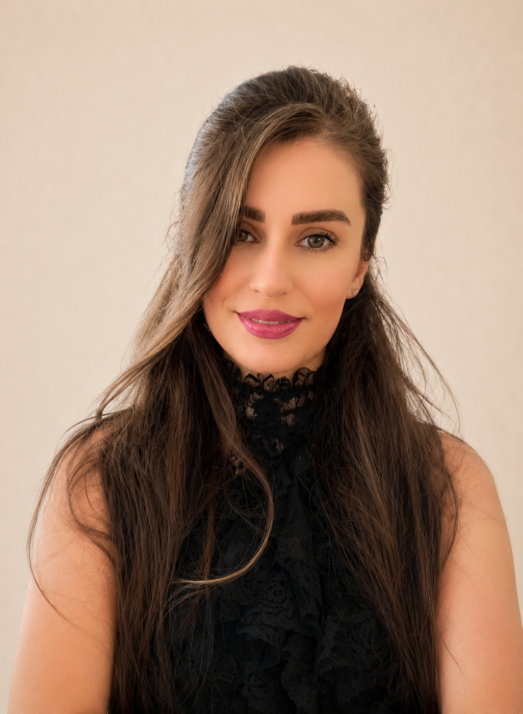 Portret van een jonge vrouw met lange, golvende bruin haar, lichte huid, groene ogen, rozen Lippen, gekleed in een zwarte kanten top, lachend tegen een beige achtergrond.