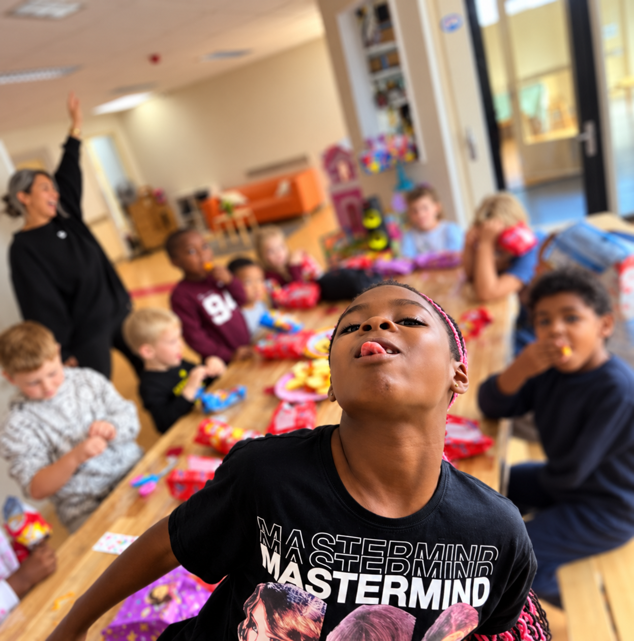 Kinderen vieren een feestje met snacks en cadeautjes aan een eettafel, een vrouw die zeert in de achtergrond.
