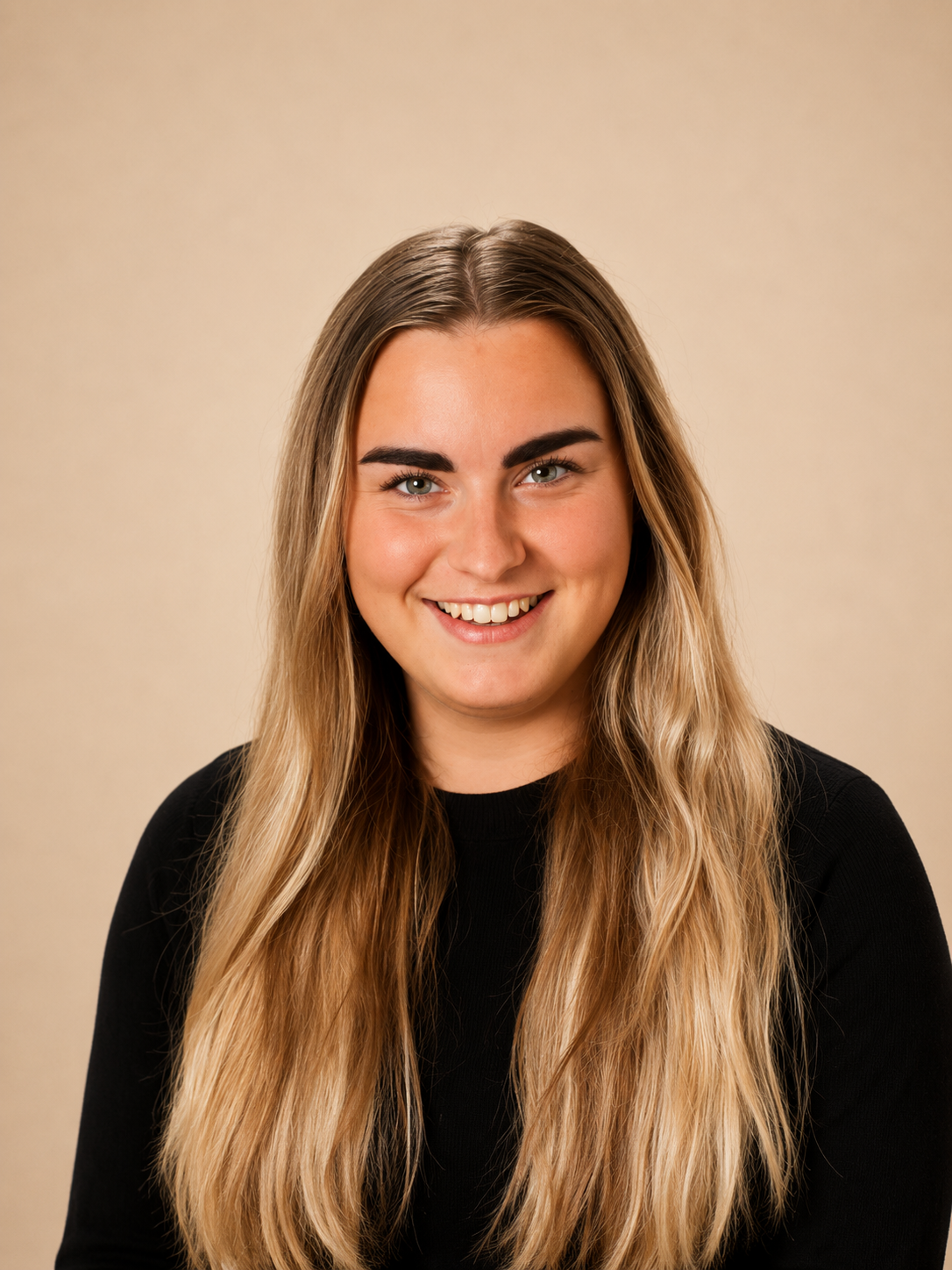 Portret van een lachende jonge vrouw met lang, golvend blond haar en een zwarte top, staand voor een beige achtergrond.