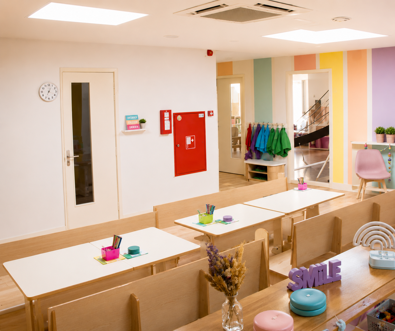 Kleurrijke en speelse kinderkamer met houten tafels en stoelen, potloden, decoraties, en kleurrijke jassen aan de kapstok. Wanden met pastelkleuren en decoratieve elementen.