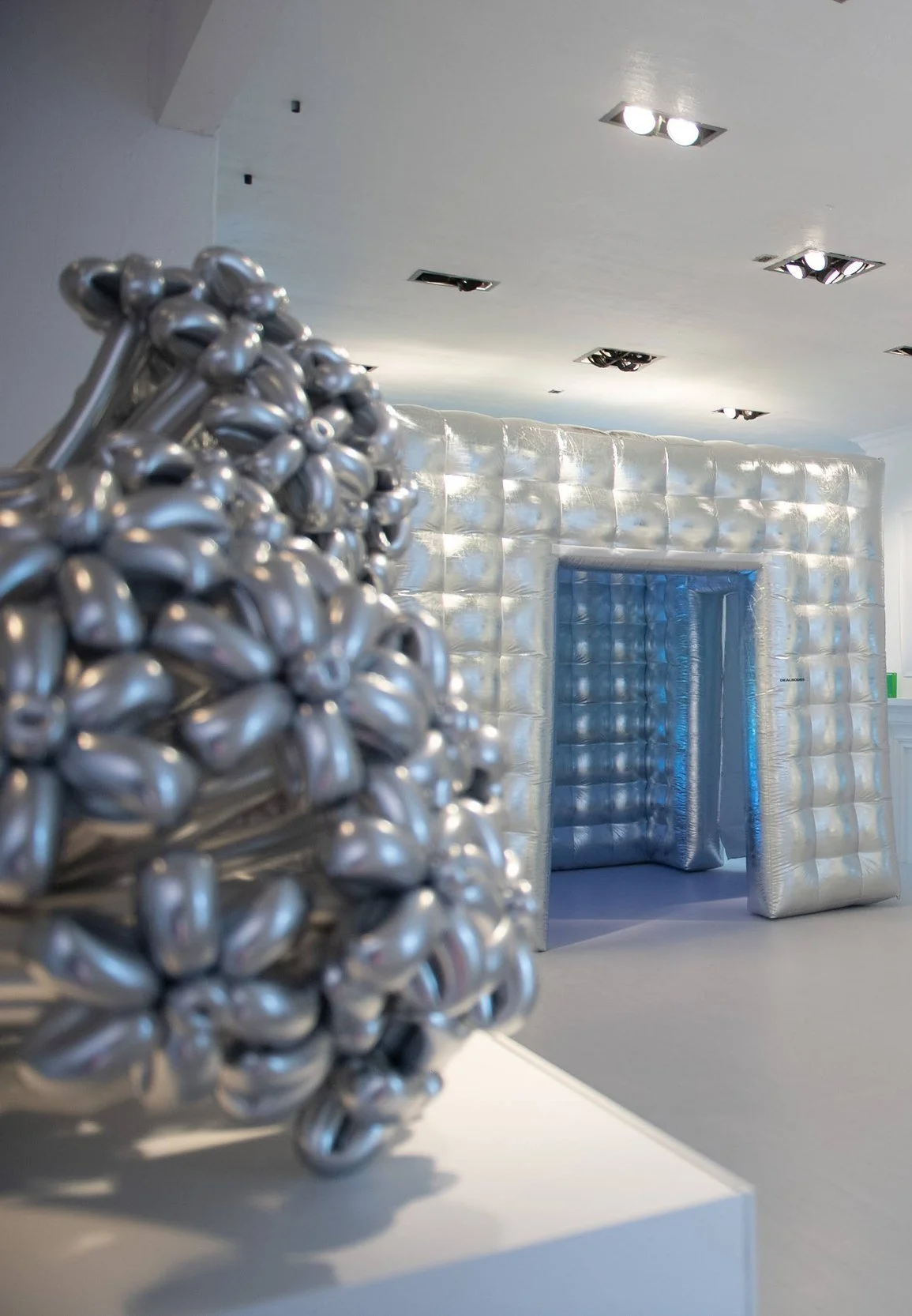 A metallic sculpture composed of numerous small, shiny, silver teardrop shapes in the foreground, with a silver inflatable or padded structure forming an archway or entrance in the background.