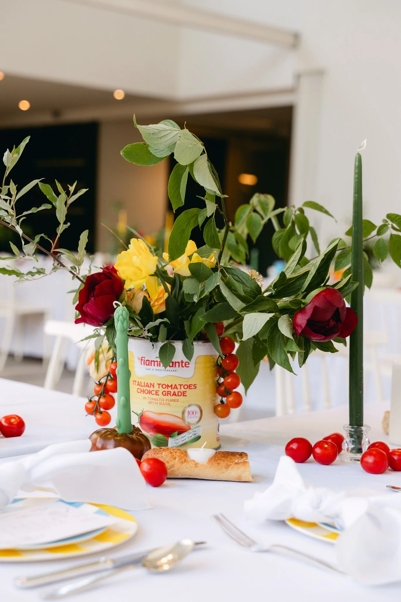 A decorated dining table with a flower arrangement, red and yellow flowers, cherry tomatoes, lighted candles, and place settings with napkins and utensils.
