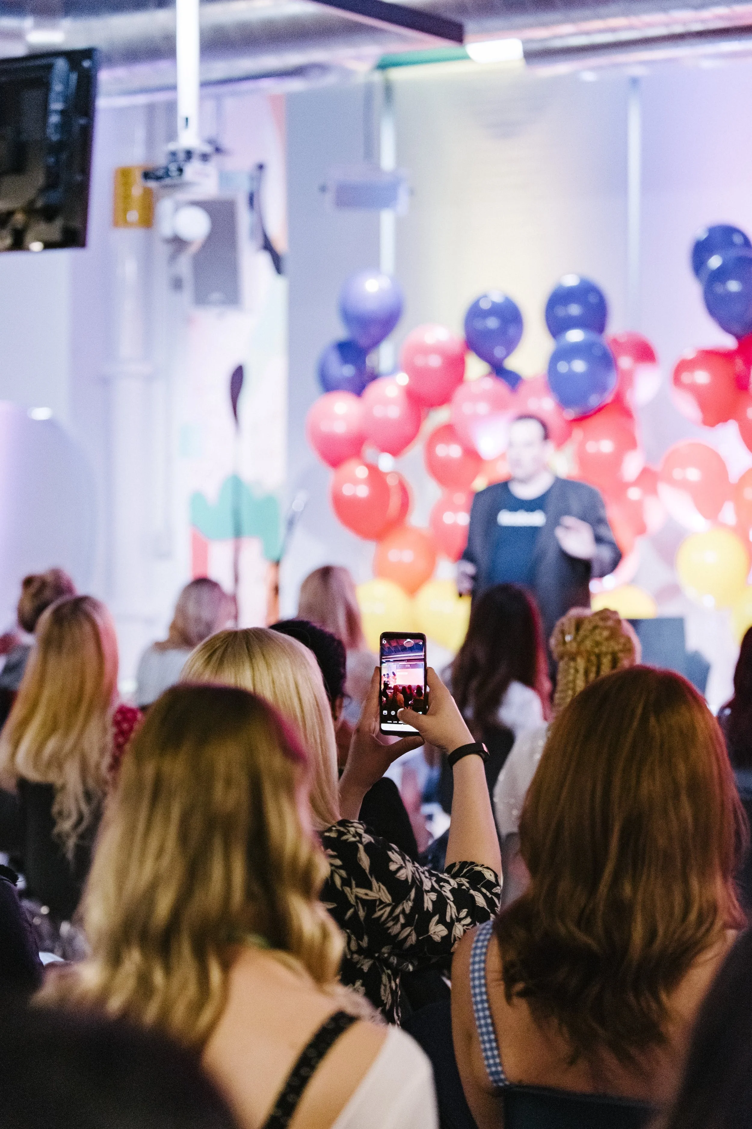 A woman taking a photo of a speaker at a conference or event with balloons in the background.