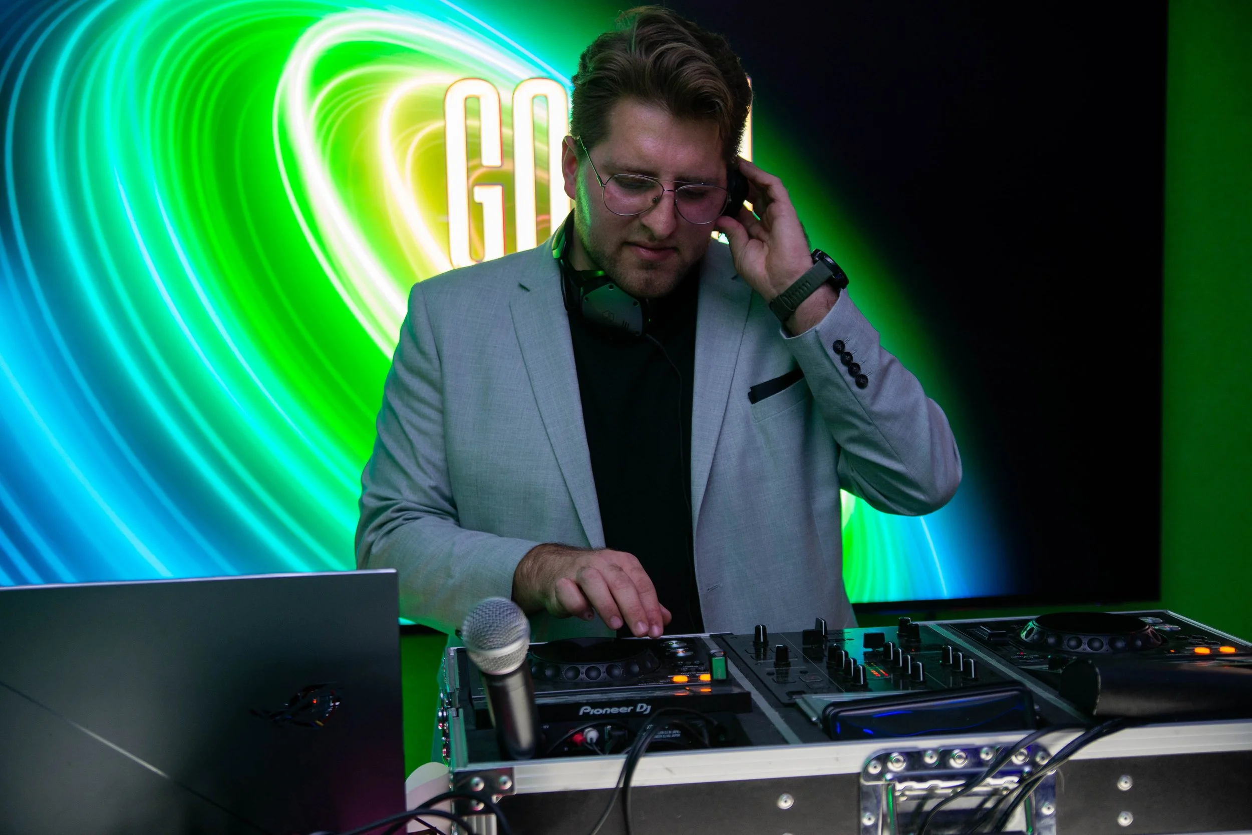 A male DJ wearing glasses and a gray suit jacket with a black shirt, using DJ equipment with headphones around his neck, in front of a green and blue swirl light background.