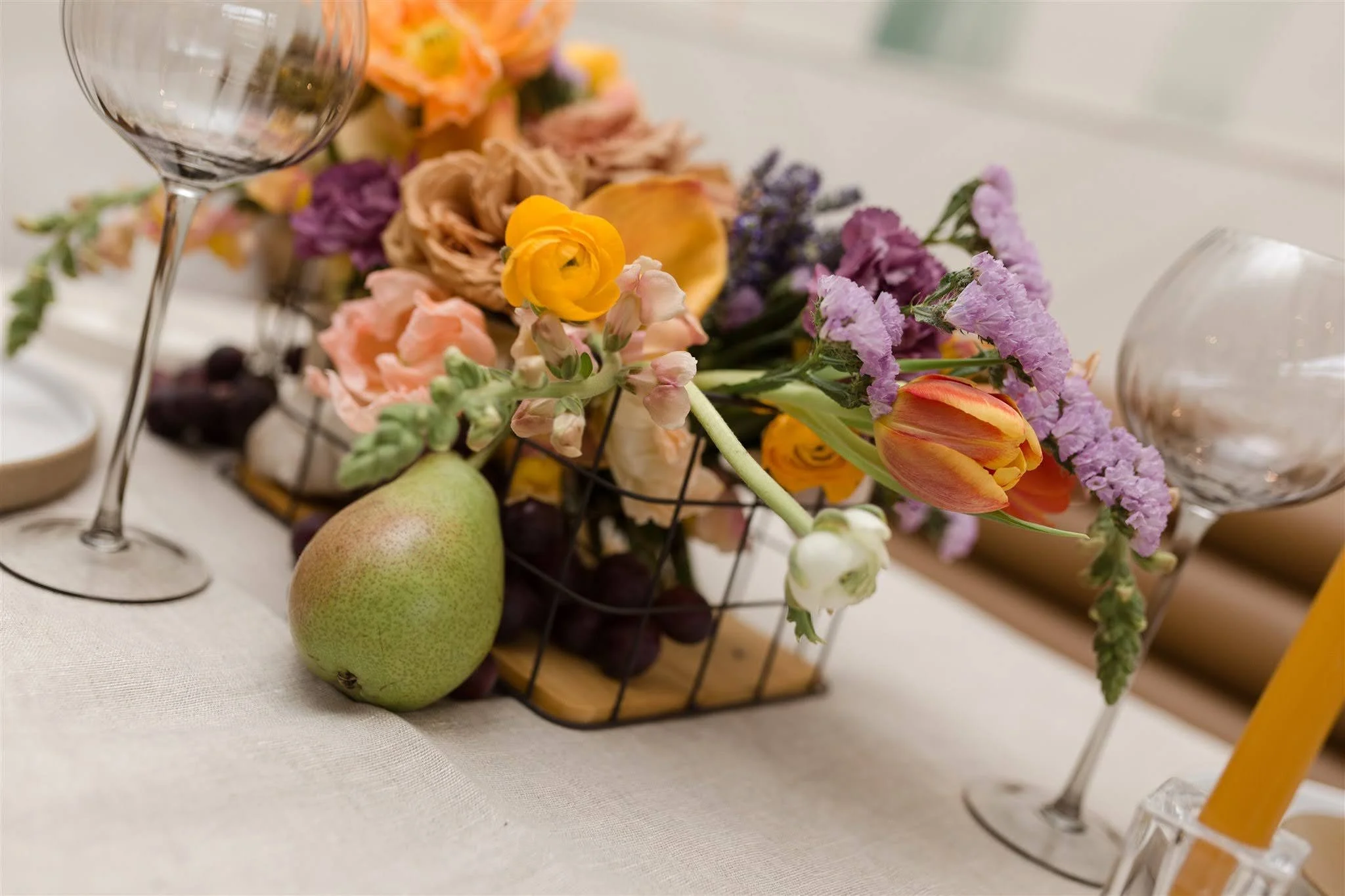 Arrangement of colorful flowers in a wire basket, a green pear, empty wine glasses, and a yellow candle on a white tablecloth.