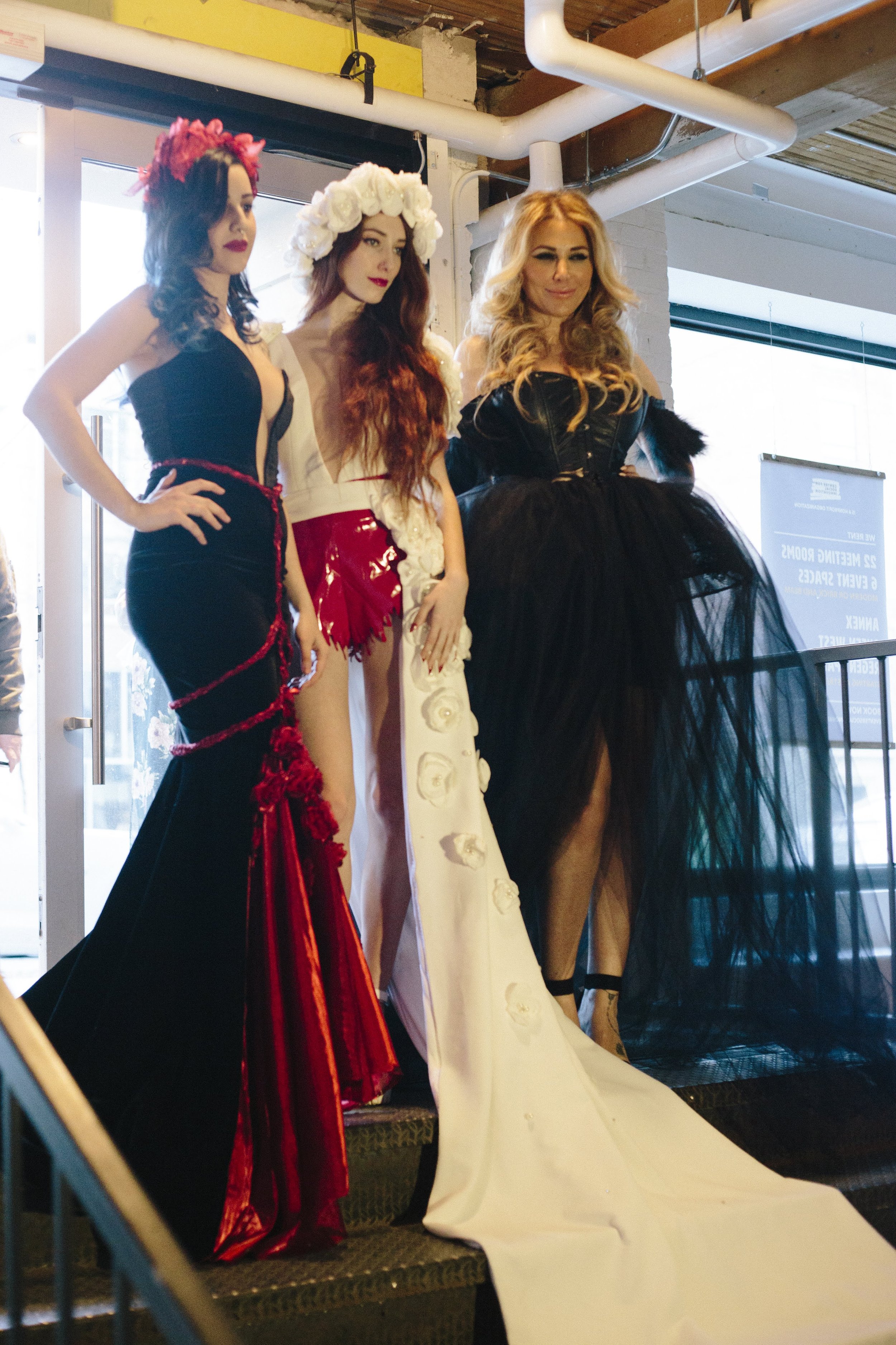 Three women in elaborate dresses standing on a staircase at a fashion event.