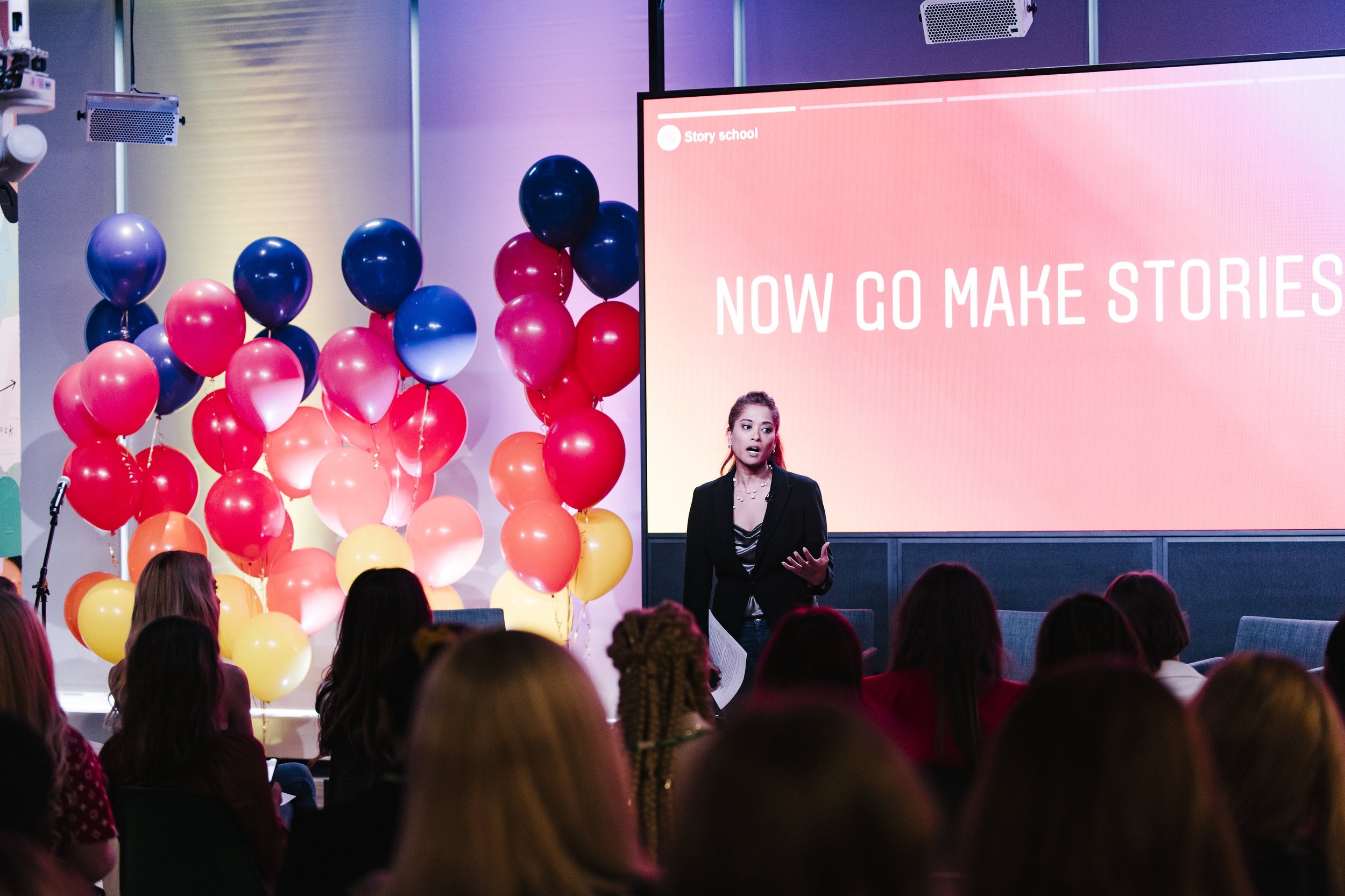 A woman giving a presentation in front of a large screen that reads 'Now go make stories.' Patterned balloons in pink, blue, yellow, and orange decorate the left side of the scene. Audience members are seated and listening.
