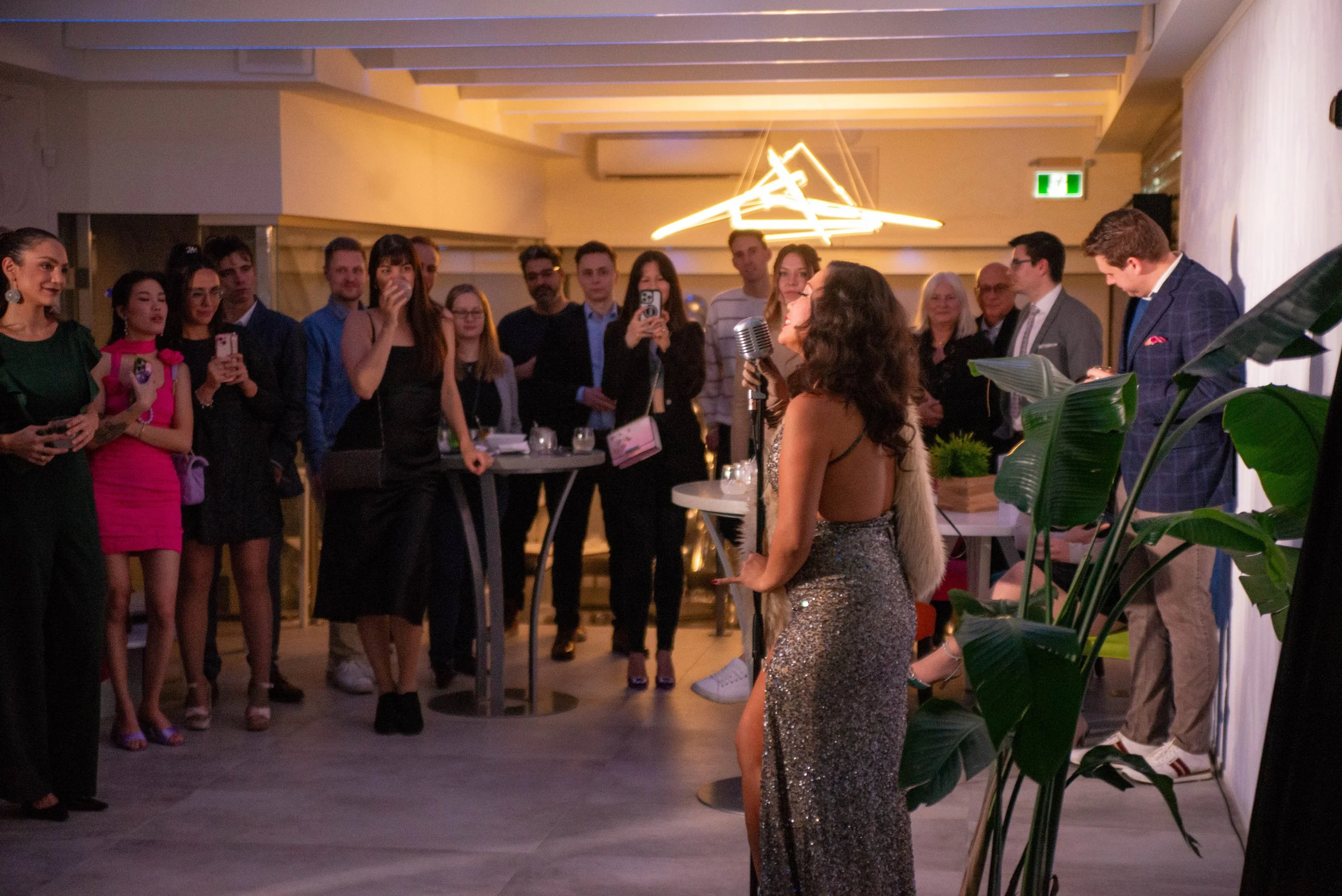 A woman in a shimmering silver dress is performing with a vintage microphone at a party or event, with an audience of well-dressed people watching and taking photos, in a decorated indoor setting with modern lighting and wall decor.