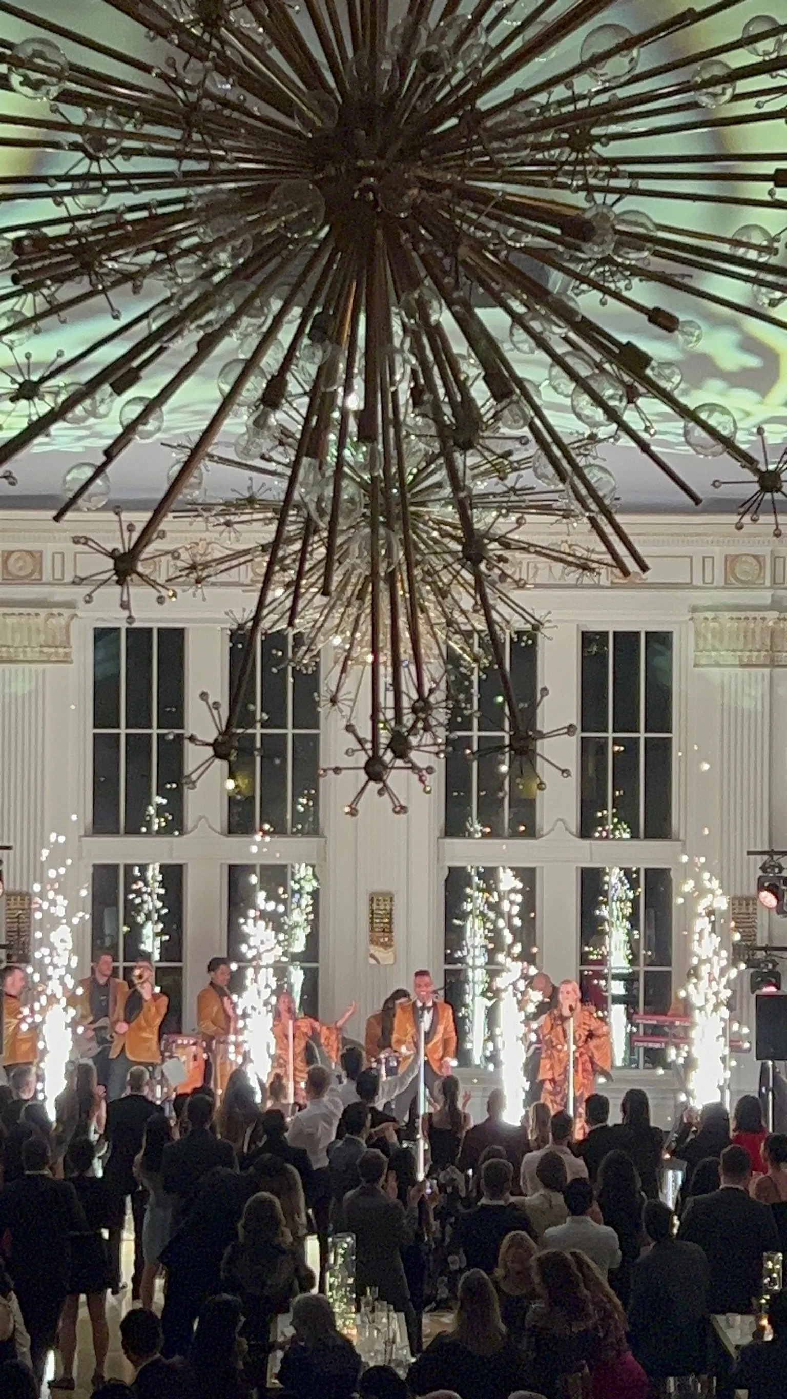 A live musical performance at an elegant indoor event with a large audience, golden-clad performers on stage, bright sparklers, and an elaborate modern chandelier hanging from the ceiling.