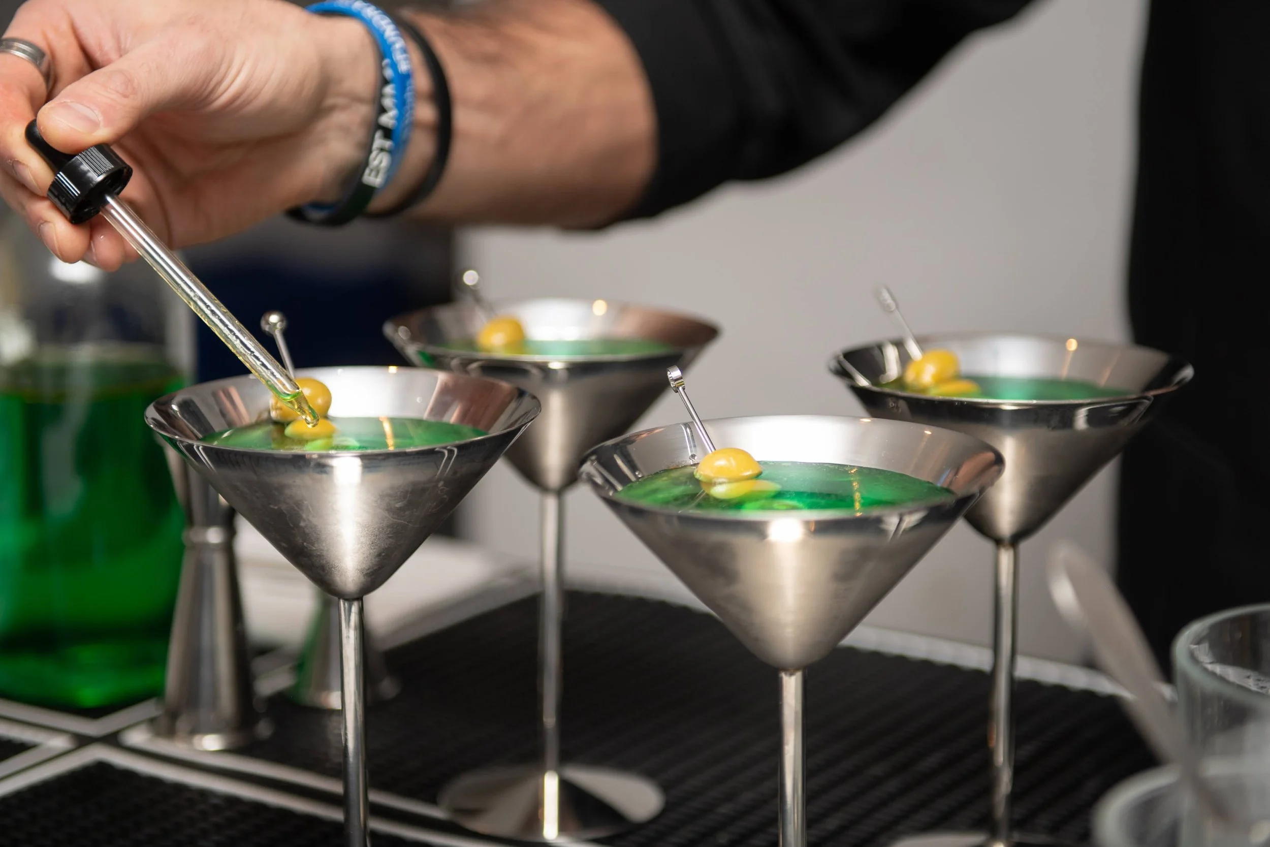 A person using a dropper to add liquid to a green cocktail garnished with olives in martini glasses.