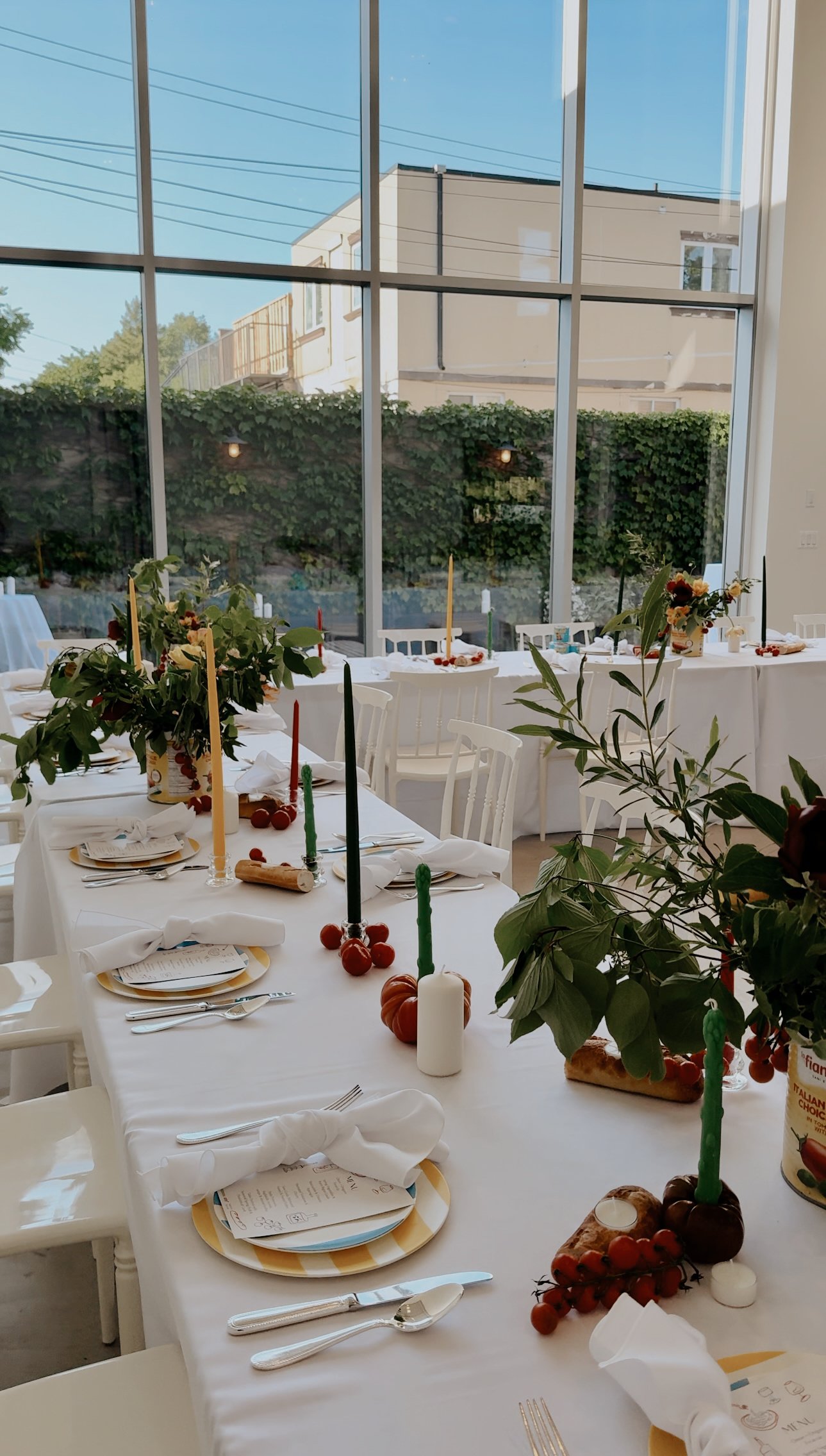 A decorated dining table set for a celebration inside a room with large glass windows, sunlight streaming in, and outdoor greenery visible through the windows.