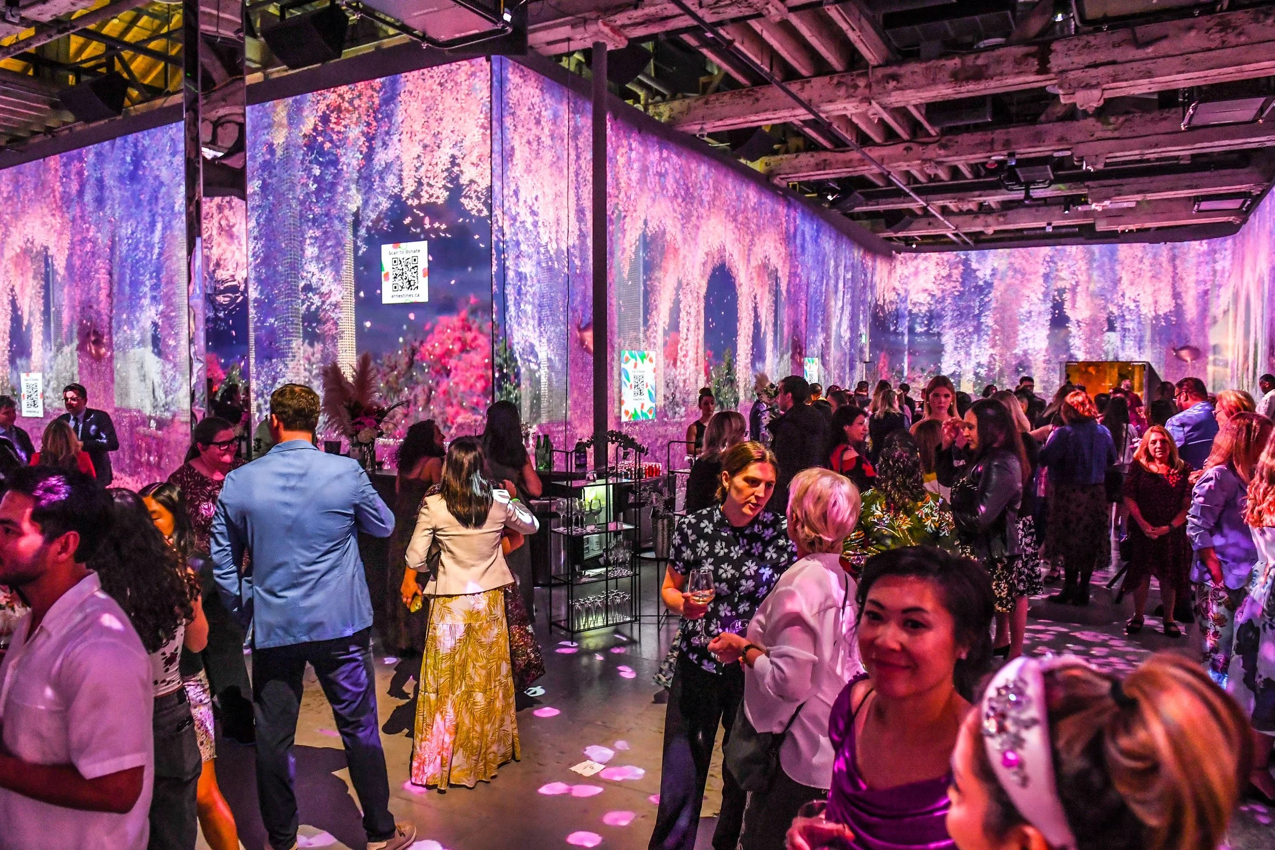 People attending a colorful indoor event with vibrant floral and nature-themed digital projections on walls, some holding drinks, and socializing in groups.