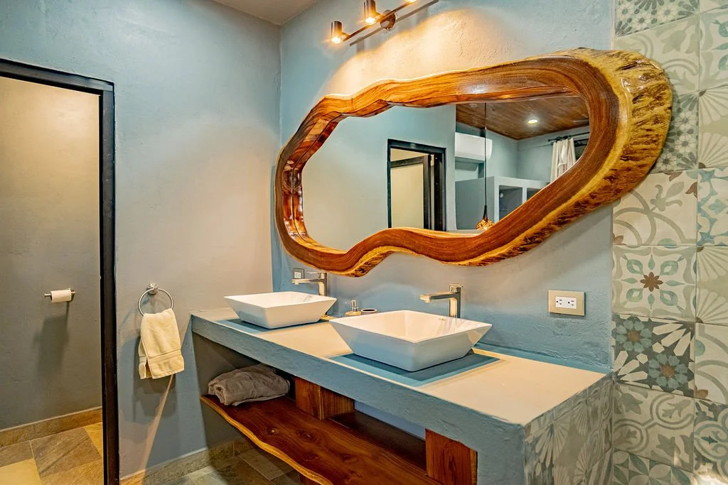 Bathroom with two rectangular vessel sinks on a floating stone counter, large irregular wooden mirror, light blue walls, patterned tiles, towel ring with hand towel, and a door.