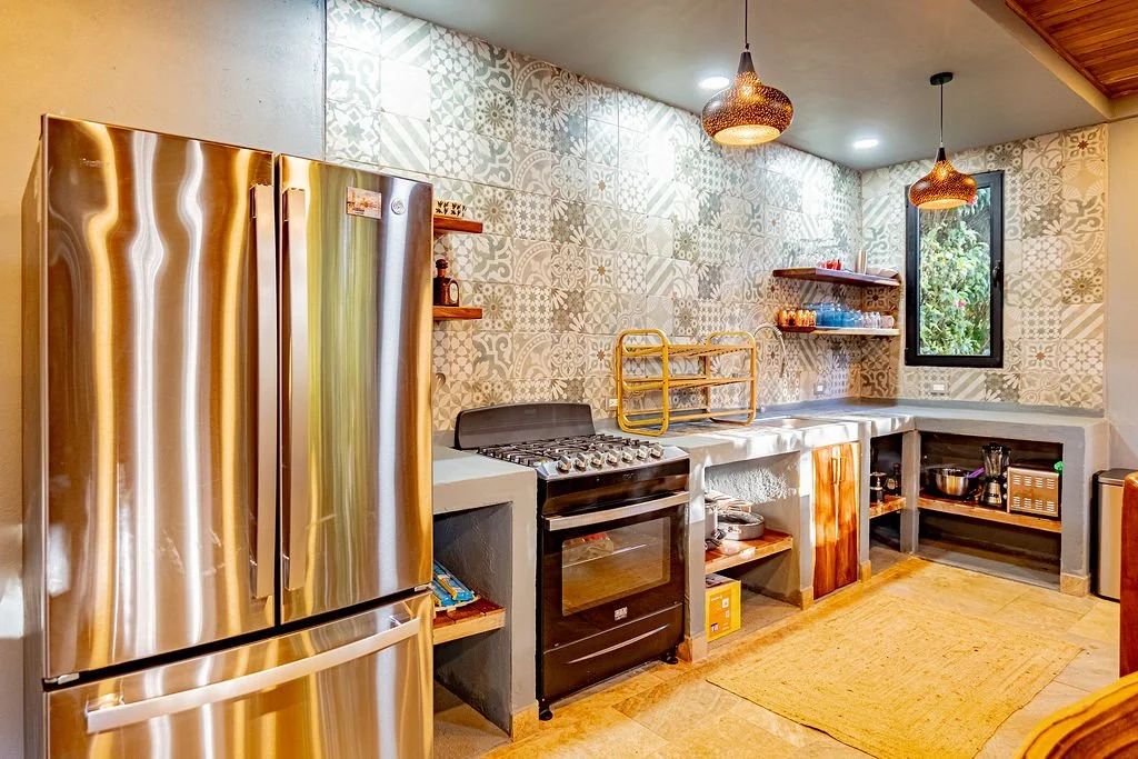 Kitchen with stainless steel refrigerator, stove, open shelving, patterned tile wall, window, and two hanging pendant lights.