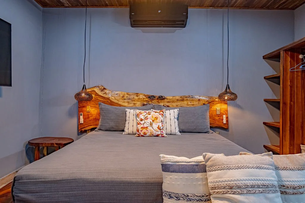 A cozy bedroom with a wooden headboard, gray bedding, decorative pillows, hanging pendant lights, a side table, and an open wooden shelving unit.