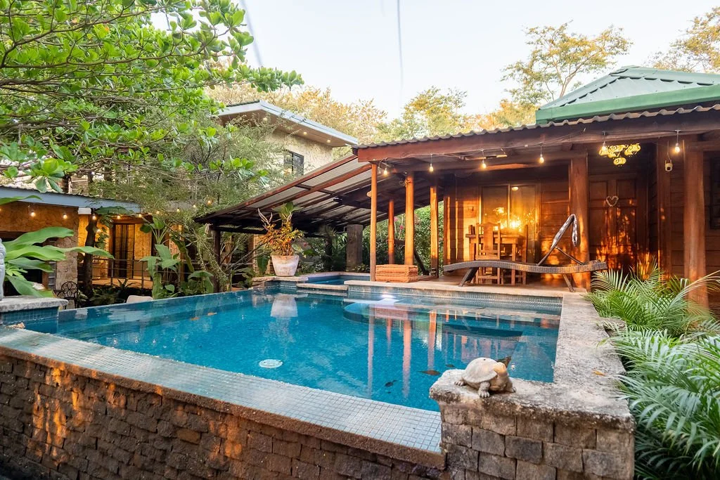 A backyard scene featuring a swimming pool with water, surrounded by greenery and trees, with a wooden house and outdoor seating area illuminated by string lights in the background.