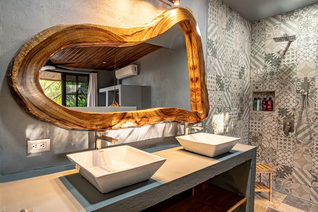 Bathroom with a large irregular-shaped wooden framed mirror, two vessel sinks, patterned tile shower area, and a small wooden stool under the sinks.