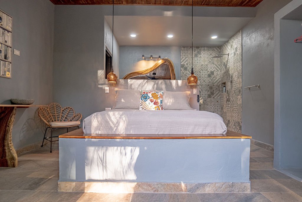 A modern bedroom with a bed, decorative pillows, a pendant light, and a walk-in shower in the background.