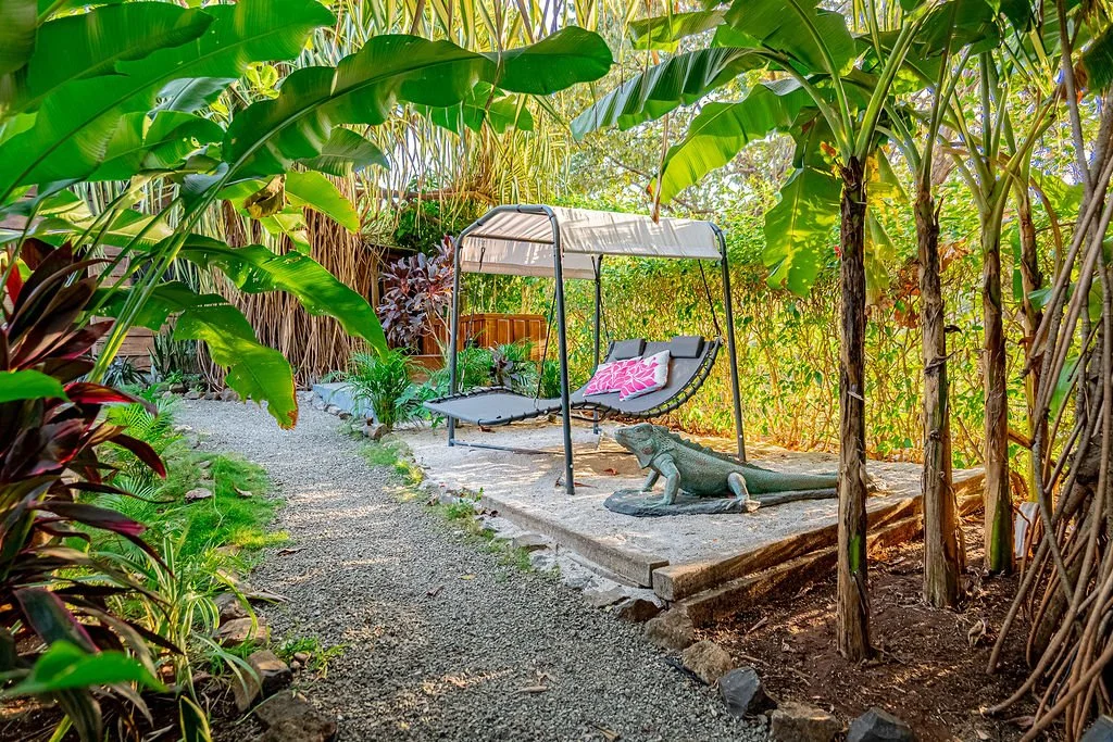 A garden scene with a gravel pathway, tropical plants, and a small patio area featuring a hanging lounge chair with cushions and pillows, surrounded by lush greenery and a stone crocodile sculpture.