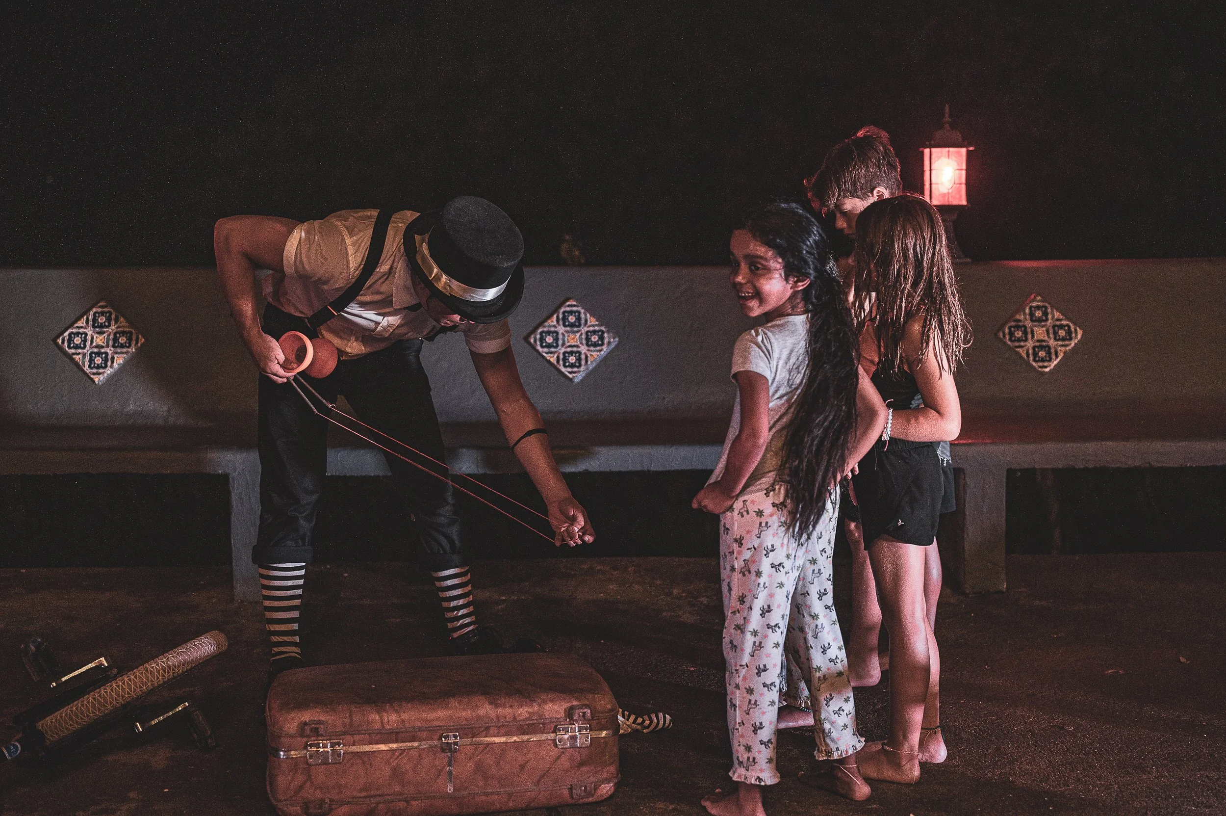 A clown performing magic tricks for three children on a dimly lit stage. The children are smiling and watching intently. The clown is dressed in a hat, suspenders, and striped socks, holding a magic ring. There is a suitcase and a juggling pin on the