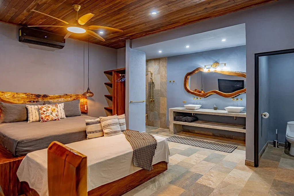 A rustic-style hotel room with a wooden ceiling, ceiling fan, and split air conditioning. It features a large bed with multiple pillows, a wooden bookshelf, a unique mirror-shaped sink area with two vessel sinks, a TV, and a bathroom with a toilet vi