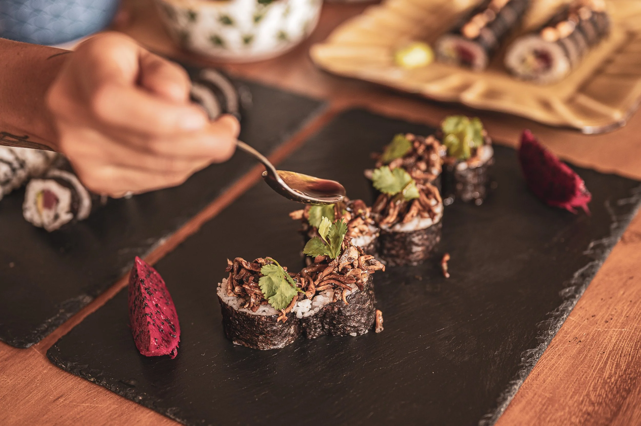 A hand drizzle dark sauce over sushi rolls topped with shredded meat and cilantro, on a black slate serving platter, with additional sushi and red pickled ginger in the background.