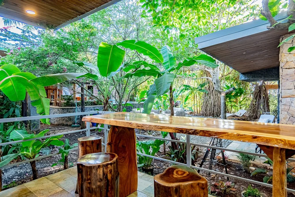 Outdoor seating area with a wooden table and matching stools, surrounded by lush green plants and trees, with a house or building in the background.