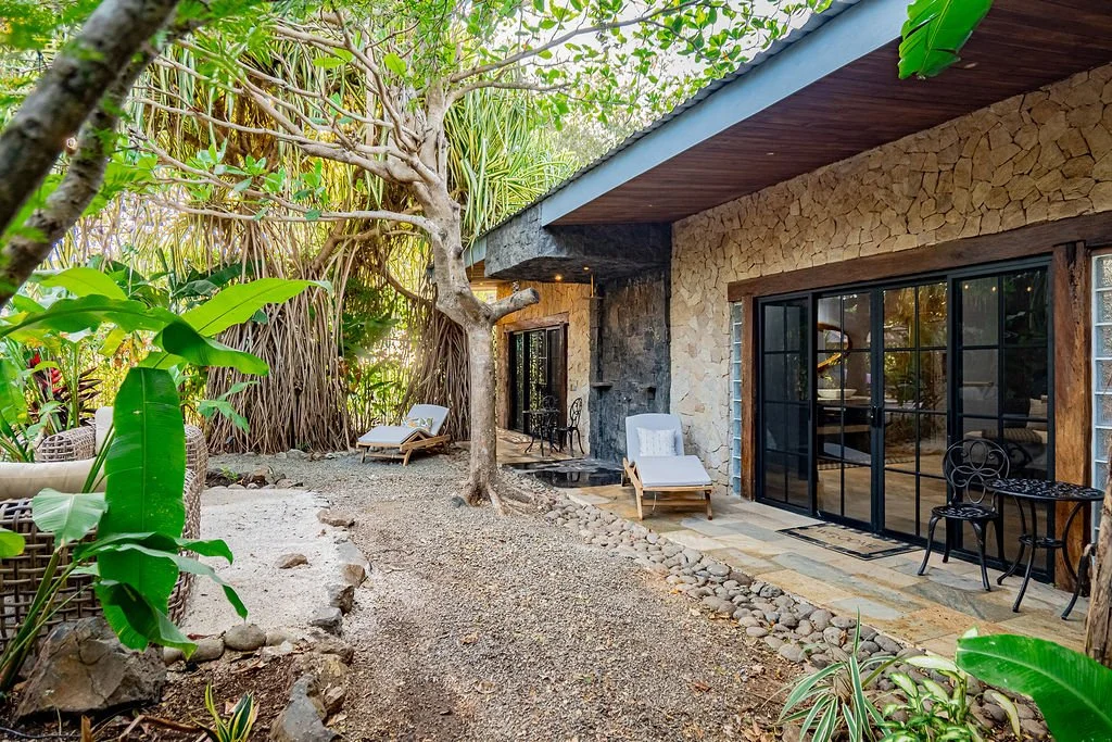 An outdoor patio area next to a house with a stone exterior and large glass sliding doors. There are lounge chairs, potted plants, and a tree in a natural, lush garden setting.