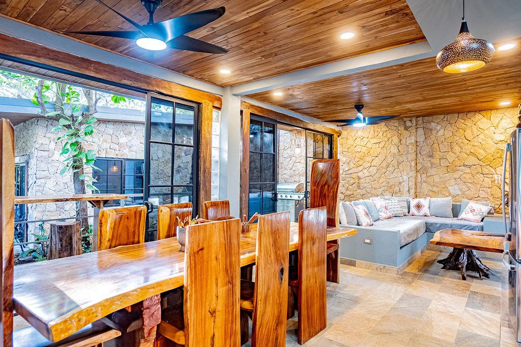 A dining area with a large wooden table and high-backed wooden chairs, a cozy corner with a built-in couch with pillows, stone wall, wooden ceiling with ceiling fans, large windows, and decorative lighting.