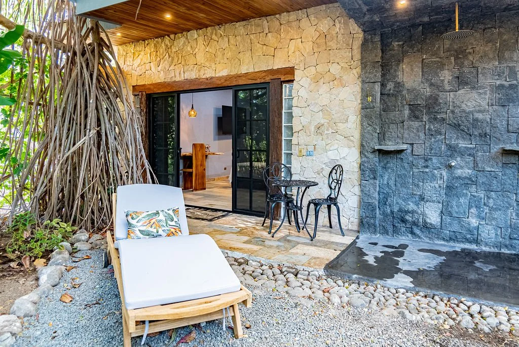 Outdoor patio area with a lounge chair, small metal table with two chairs, and a rock wall shower with a rain showerhead. Behind, there are sliding glass doors leading inside the house, which has stone and wood exterior walls.