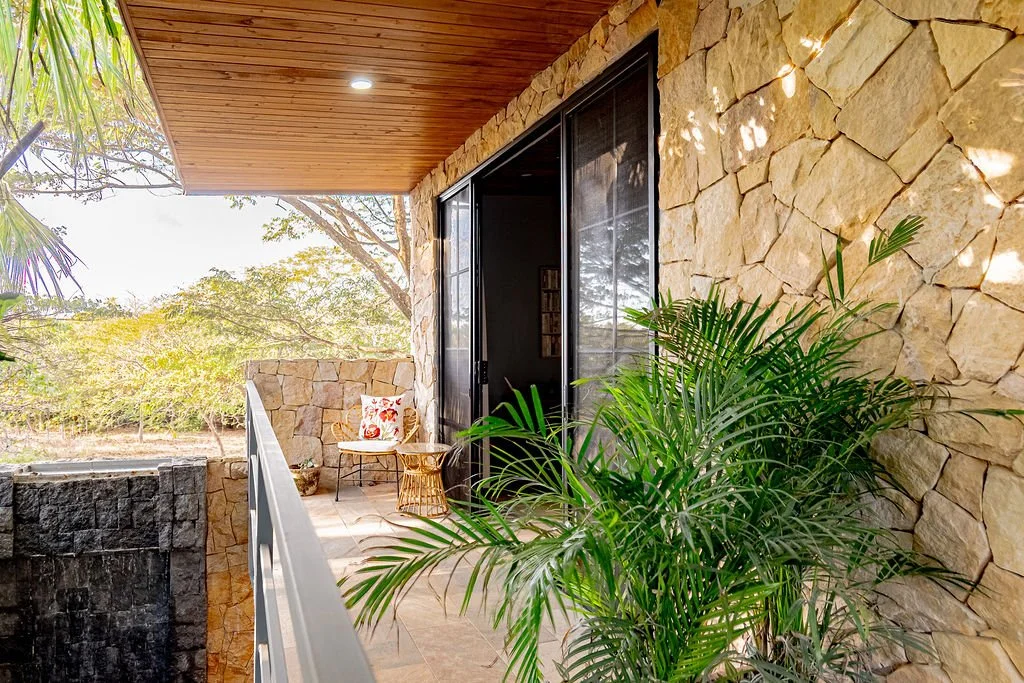 A small outdoor balcony with stone walls and a wooden ceiling, featuring a green potted plant, a small round wicker table, and a chair with a floral pillow. Glass sliding doors lead inside, with a view of lush trees and foliage in the background.