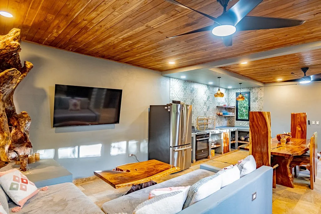 Open-concept living room and kitchen with wooden ceiling, gray walls, wall-mounted TV, gray sofa, wooden coffee table, stainless steel refrigerator, stove, open kitchen shelves, dining table with six wooden chairs, pendant lighting, window with green