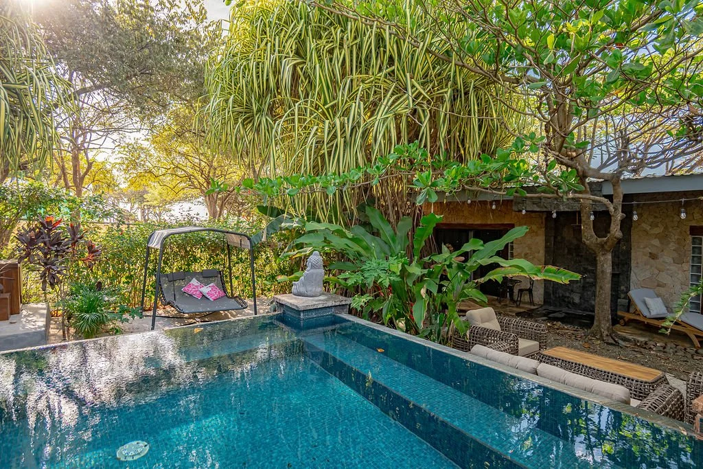 A backyard with a swimming pool surrounded by lush green plants and trees, with outdoor seating including a swing, chairs, and a small table, and a stone building in the background.