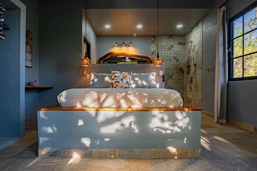 A cozy bedroom with a large bed, a decorative pillow, and two pendant lights hanging on either side. Sunlight filters through a window, creating patterns on the bed and wall. The room features a modern rustic decor with tiles on the wall and wooden f