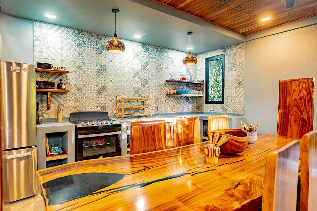 A kitchen with patterned tile backsplash, wooden dining table, and chairs, stainless steel refrigerator, stove, open shelving, and a small window.