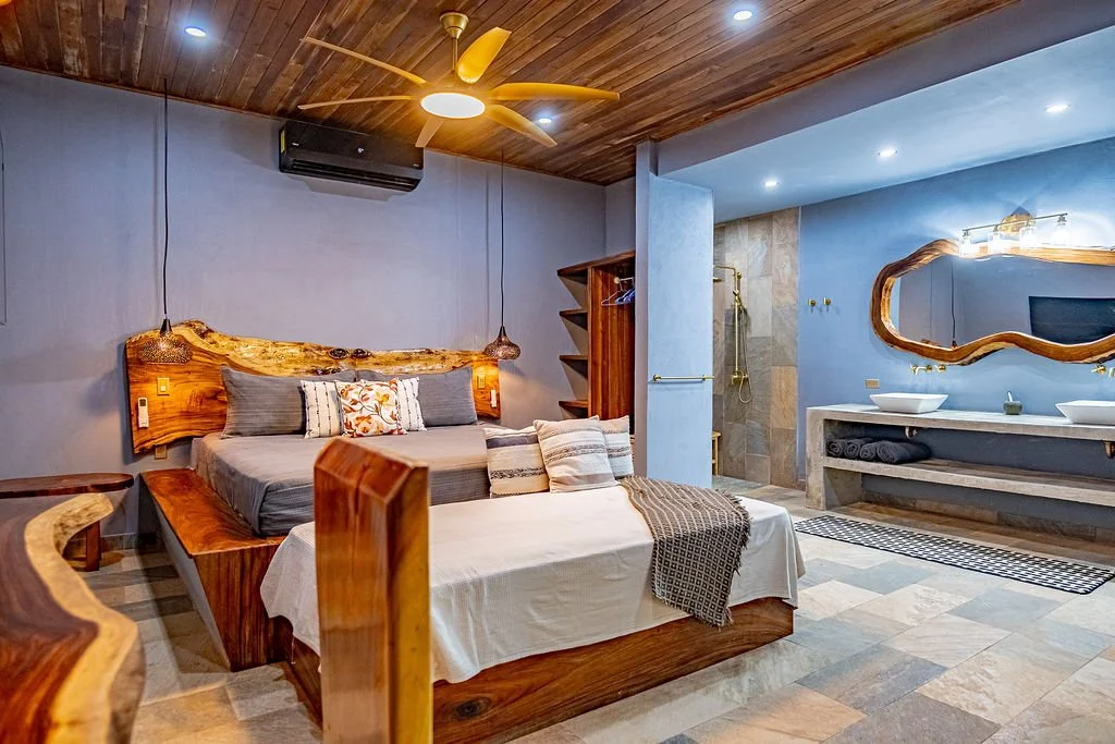 A modern bedroom with a wooden bed, gray bedding, and decorative pillows, adjacent to an open bathroom with sinks, a large mirror, and a walk-in shower.