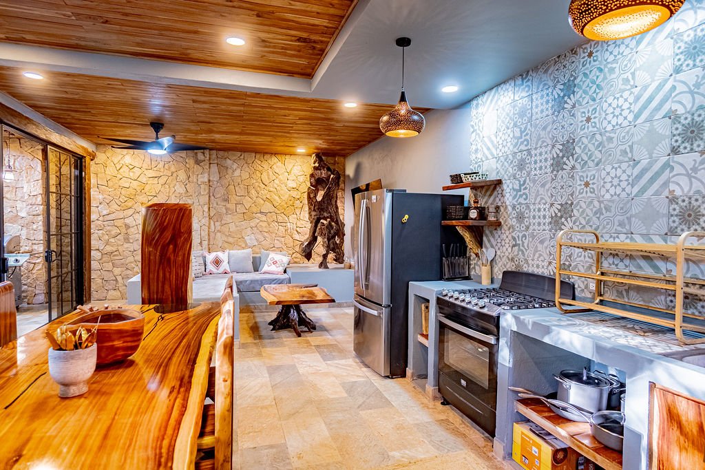 A cozy kitchen and living room with wood accents, stone walls, and modern appliances, including a stainless steel refrigerator and a stove with an oven, decorated with patterned tile walls and warm lighting.