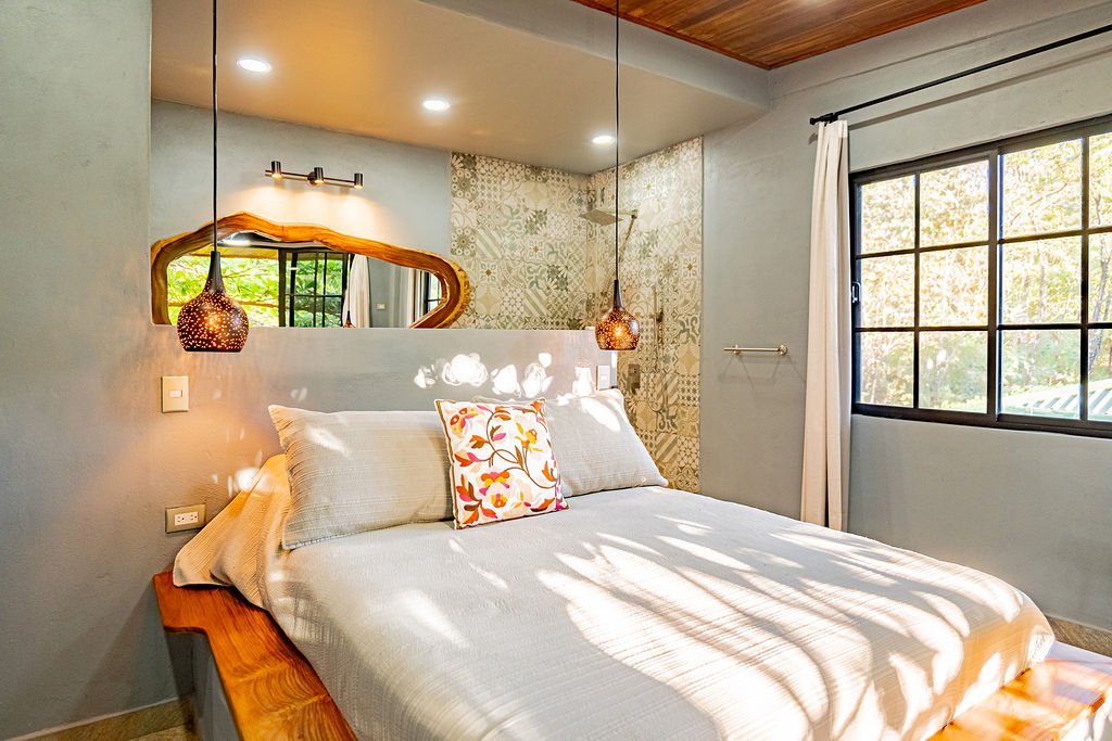 A bedroom with a bed, decorative pillows, hanging lights, a mirror, window with curtains, and sunlight casting shadows.