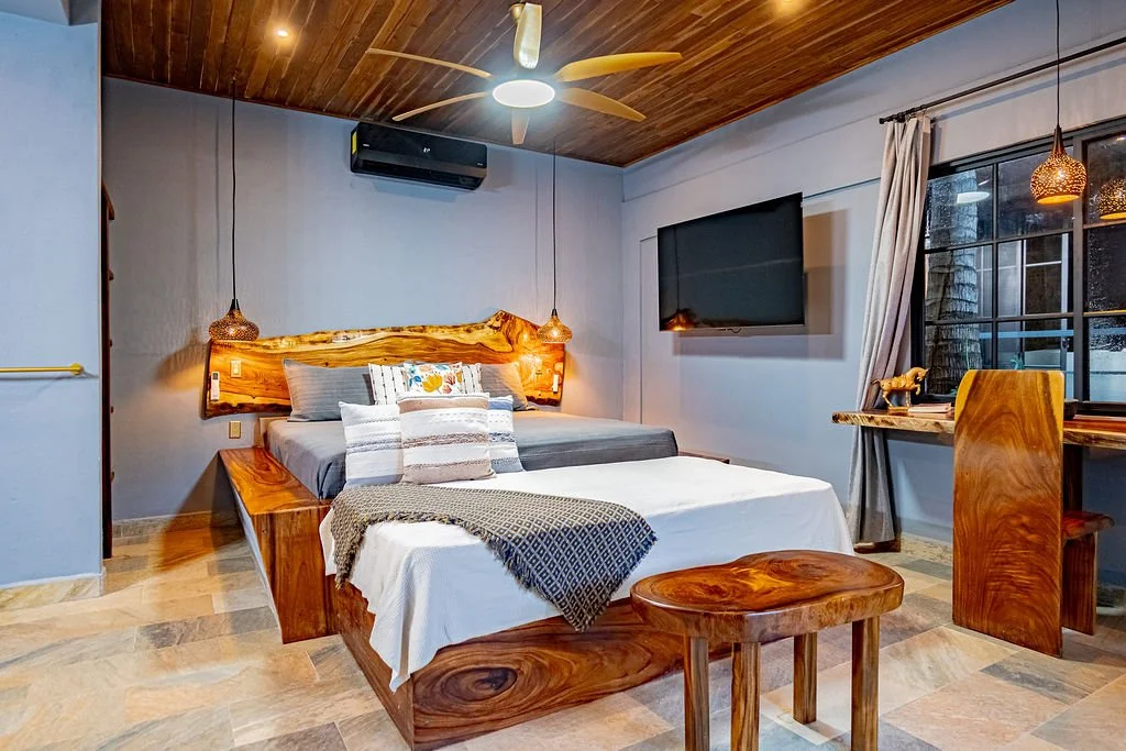 A cozy bedroom with a wooden bed, gray bedding, and decorative pillows. There is a wooden headboard and accents, a wall-mounted TV, a window with curtains, and hanging pendant lights.