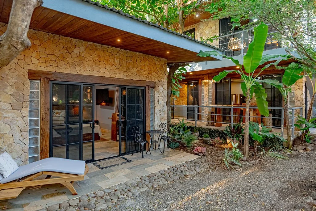 Exterior view of a stone house with a patio featuring black sliding glass doors, outdoor seating, lush greenery, and potted plants, with multiple levels and balcony railings.