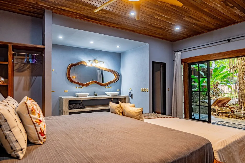 A bedroom with a wooden ceiling, a large bed with beige bedding and pillows, a sliding glass door opening to a patio with greenery, and a double vanity with two vessel sinks and a decorative mirror.